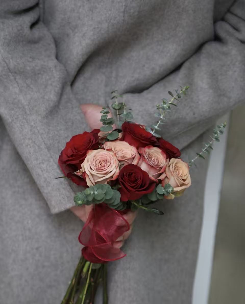 A close-up of a hand holding a small bouquet of dried roses in deep red, dusty pink, and peach tones, accented with green eucalyptus stems, against a person's gray wool coat.