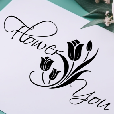 Black cursive script reading 'Flower You' with tulip flower silhouettes, printed on a white card or paper over a teal surface with small green leaves and pale flowers in the corner.