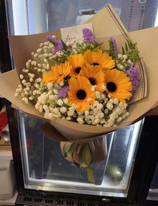 A floral bouquet wrapped in brown kraft paper, featuring bright yellow-orange gerbera daisies, white baby's breath, and small purple flowers, held in front of a glass display case.