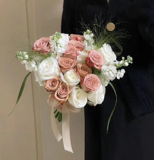 A bouquet of pink and white roses with white filler flowers and green stems, held by a person wearing a black cardigan against a beige indoor background.
