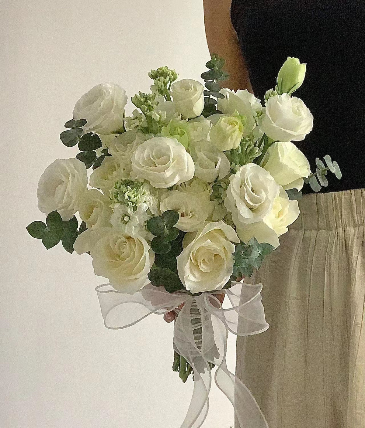 A bouquet of white roses and green foliage tied with a sheer white ribbon, held by a person wearing a black top and beige skirt against a plain light background.