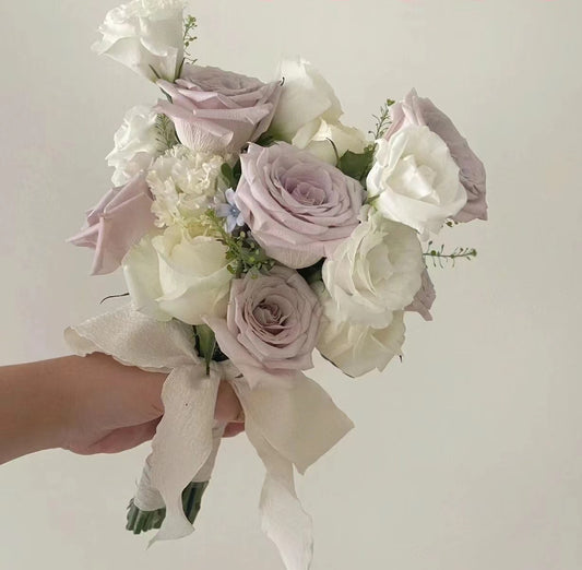 A hand holding a bouquet of pale lavender and white roses with a cream ribbon, set against a plain off-white background.