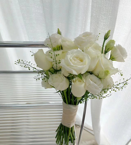 A bouquet of white roses and lisianthus flowers with green stems, wrapped in a cream paper band, set against a background of white sheer curtains and window blinds.