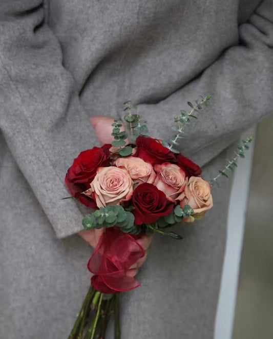 A close-up of a hand holding a small bouquet of dried roses in deep red, dusty pink, and peach tones, accented with green eucalyptus stems, against a person's gray wool coat.