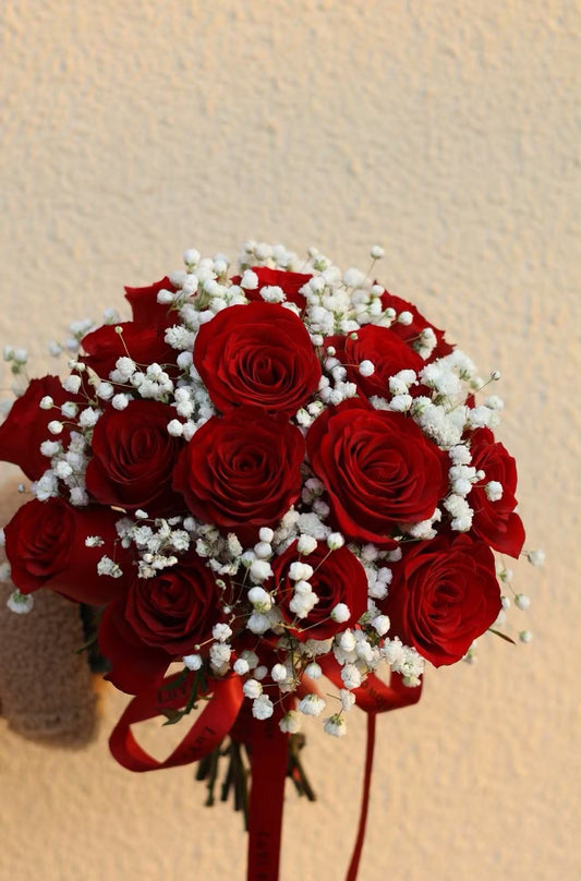 A bouquet of red roses and white baby's breath flowers tied with a red ribbon, set against a textured beige wall.