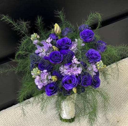 A vibrant floral bouquet featuring deep purple roses, light purple lisianthus, and green dill, arranged in a white-wrapped vase on a textured beige surface.