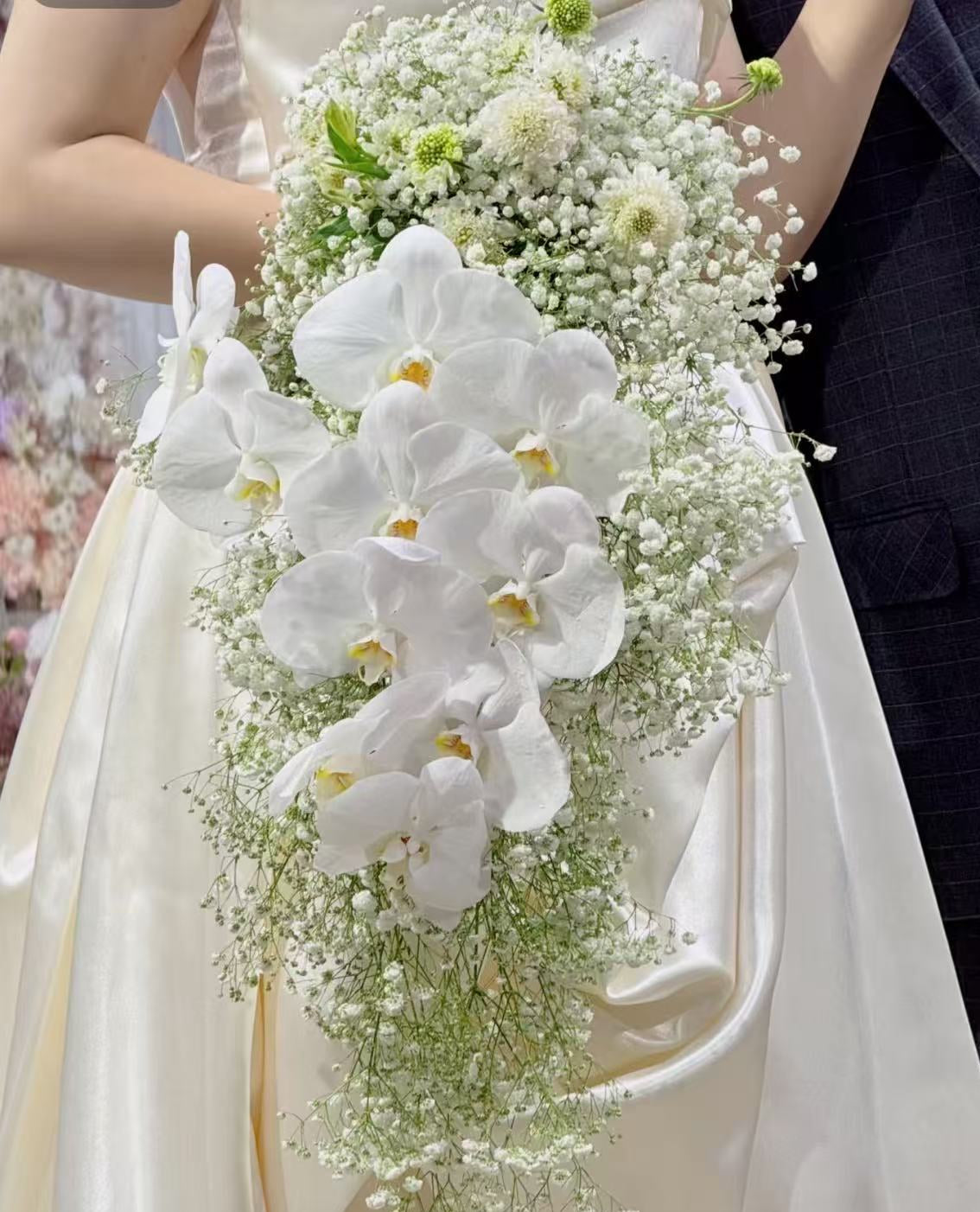 A bridal bouquet featuring large white orchids and delicate white baby's breath, held by a bride in a cream satin wedding dress next to a dark navy suit.