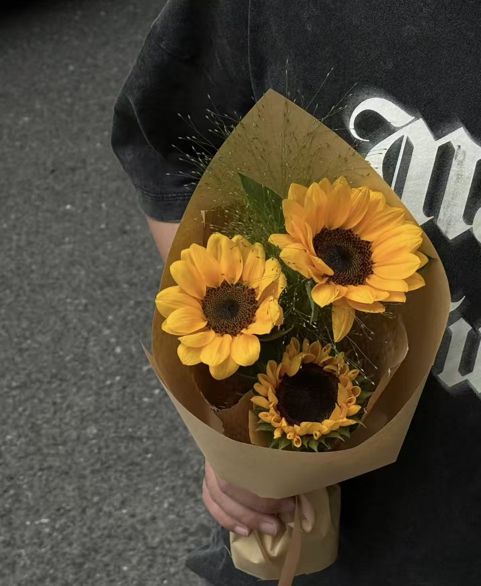 A bouquet of three yellow sunflowers wrapped in brown kraft paper, held by a person wearing a black graphic t-shirt against a gray asphalt background.