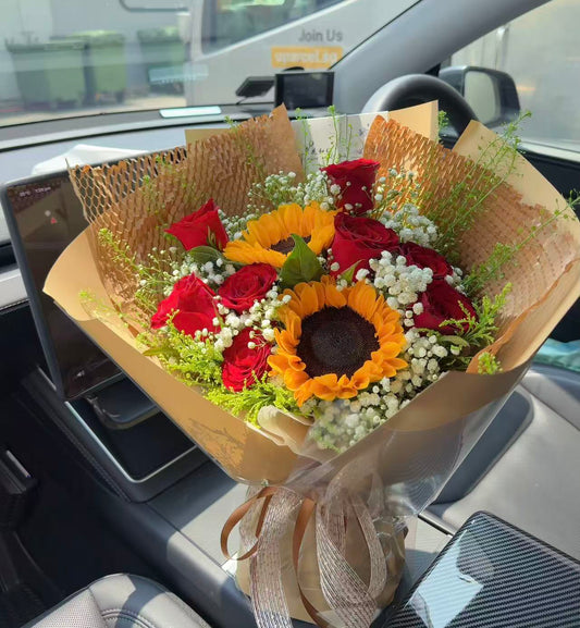 A floral bouquet arranged in a car interior, featuring red roses, a large yellow sunflower, white baby's breath, and green foliage wrapped in tan paper and ribbon.